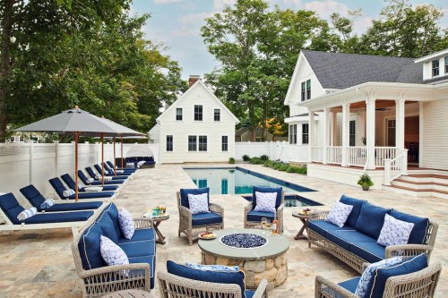 A stylish outdoor patio with a pool, lounge chairs, and a fire pit, surrounded by trees and a white house.