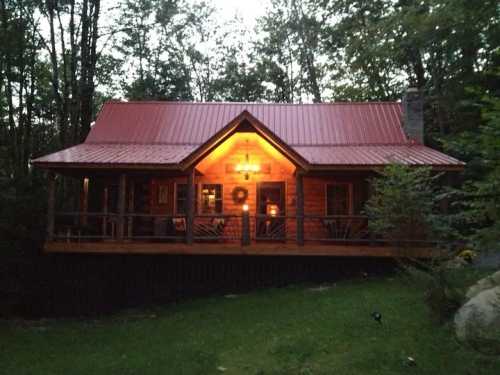 A cozy wooden cabin with a red metal roof, warmly lit at dusk, surrounded by trees and a grassy area.