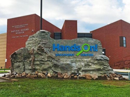 Sign for Hands-On Discovery Center in front of a natural history museum, featuring a rock formation and dinosaur fossil.
