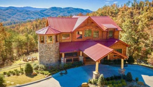 A large wooden house with a red roof, surrounded by trees and mountains in the background, on a sunny day.