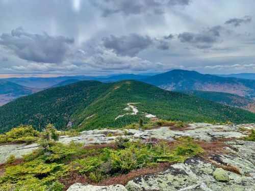 A panoramic view of green mountains under a cloudy sky, with rocky foreground and autumn foliage in the distance.