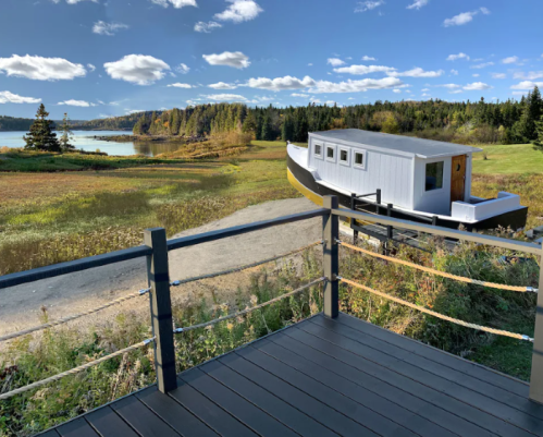 A scenic view from a deck overlooking a grassy area and a unique boat-shaped house by the water under a blue sky.