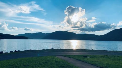A serene lake scene with mountains in the background, under a bright sky with fluffy clouds and sunlight reflecting on the water.