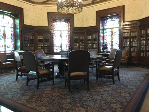 A cozy library with wooden shelves, stained glass windows, a round table, and upholstered chairs.