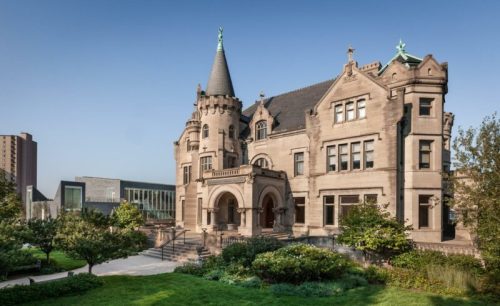A grand stone mansion with a turret and spire, surrounded by lush greenery and modern buildings in the background.