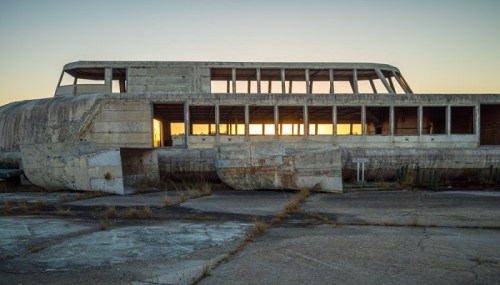 Abandoned concrete boat structure at sunset, with broken windows and overgrown surroundings.