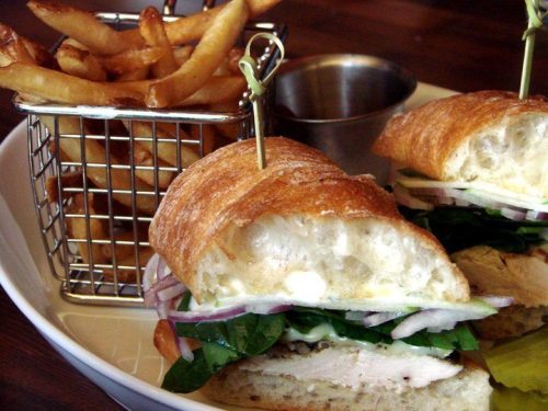 A close-up of a sandwich with chicken, spinach, and onions, served with a side of fries in a small metal basket.