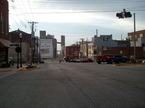 A street view of Alton, featuring buildings, parked cars, and a traffic light against a cloudy sky.