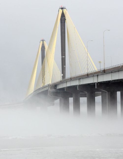A foggy scene featuring a large suspension bridge with yellow cables and tall pillars rising above the misty water.