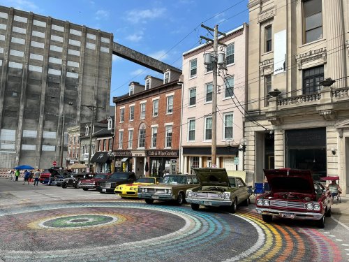 A row of classic cars parked on a colorful circular pavement in front of historic buildings.
