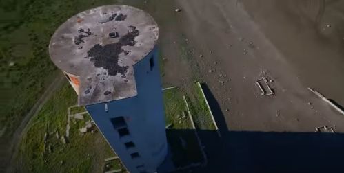 Aerial view of a tall, abandoned cylindrical tower on a barren landscape with scattered debris.