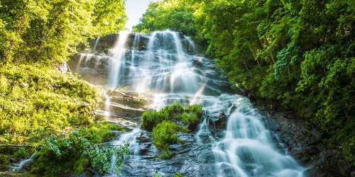 A cascading waterfall surrounded by lush green trees and vibrant foliage under bright sunlight.