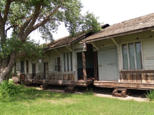 Abandoned wooden building with a porch, surrounded by greenery and trees, showing signs of weathering and neglect.