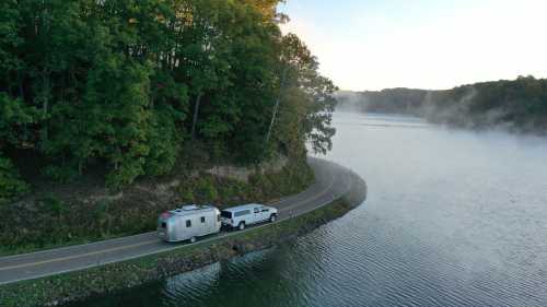 A silver Airstream trailer tows a truck along a winding road by a misty lake surrounded by trees.