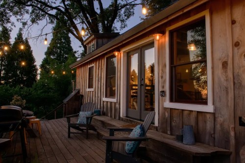Cozy wooden cabin exterior at dusk, featuring string lights, a deck, and two chairs.