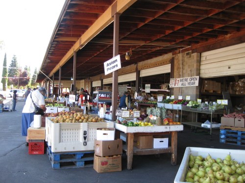 A bustling farmers market with various fruits and vegetables displayed on tables under a wooden structure.
