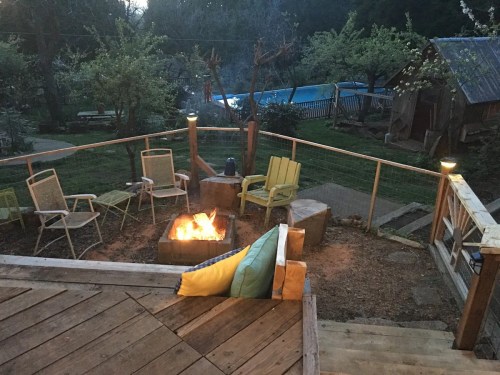 Cozy outdoor seating area with a fire pit, surrounded by trees and a pool in the background during twilight.