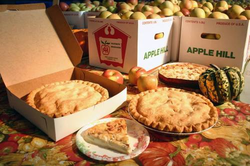 A table with apple pies, a slice of pie, and decorative gourds, surrounded by boxes of apples.