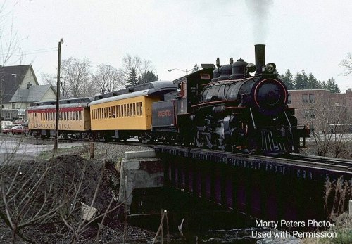 A vintage steam train crosses a bridge, with a red and yellow passenger car, surrounded by trees and buildings.