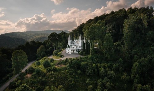 A grand white mansion with spires, nestled among lush green trees and rolling hills under a cloudy sky.