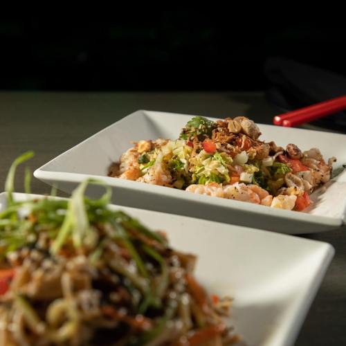 Two plates of colorful sushi dishes, one with shrimp and vegetables, the other with noodles and garnishes.