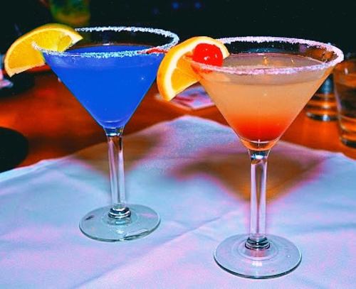Two colorful cocktails in martini glasses, one blue with a lemon slice and cherry, the other orange with a lemon slice.