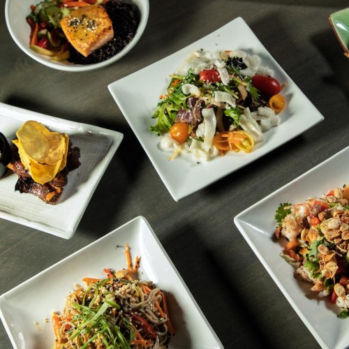 A variety of colorful dishes on white plates, including salads, noodles, and grilled salmon, arranged on a table.