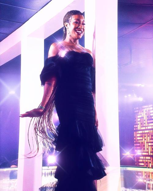 A person in a black off-shoulder dress poses confidently on a brightly lit stage with a vibrant background.