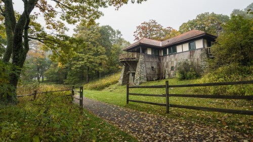 A rustic house with a wooden exterior sits among trees, surrounded by a path and a wooden fence in a serene landscape.