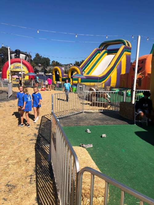 Two children in blue shirts stand near inflatable attractions at a fair, with people enjoying the activities in the background.