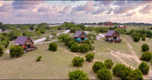 Aerial view of several rustic cabins surrounded by greenery and winding paths under a cloudy sky.