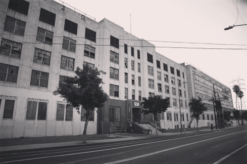 Abandoned industrial building with boarded windows, trees in front, and a street view in black and white.