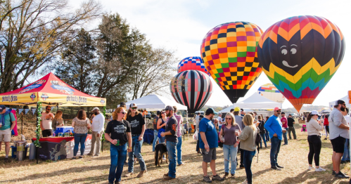 Colorful hot air balloons rise above a lively crowd at an outdoor festival with tents and trees in the background.