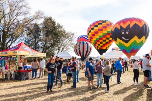 A lively outdoor festival with colorful hot air balloons and people enjoying the event amidst tents and autumn scenery.