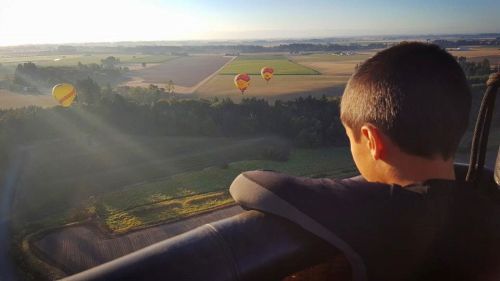 A child gazes out from a hot air balloon, overlooking fields and other balloons in the distance during sunrise.