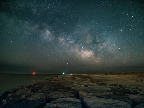 A rocky pier extends into a dark sea under a starry night sky, featuring the Milky Way and distant lights.