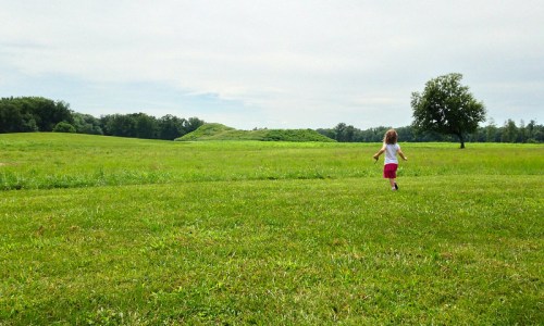 A young child runs across a grassy field towards a distant hill and a lone tree under a cloudy sky.