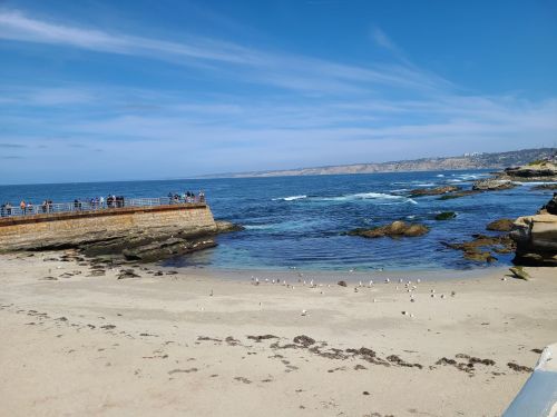 A scenic beach view with a rocky shoreline, gentle waves, and people walking along a pier under a clear blue sky.