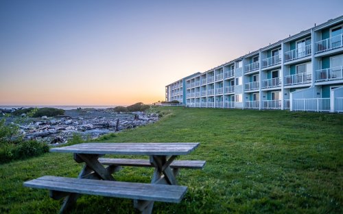 A picnic table on green grass in front of a seaside hotel at sunset, with rocky shoreline in the background.