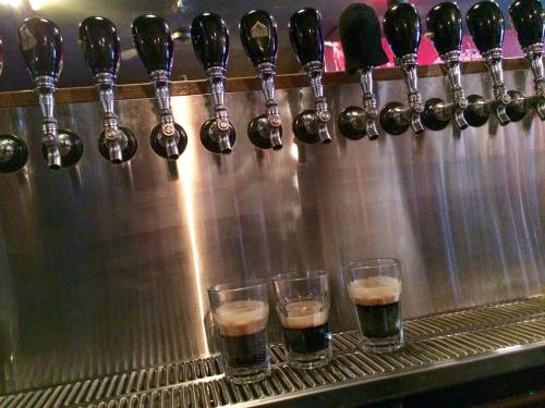 Three glasses of dark beer sit on a bar counter in front of a row of beer taps.
