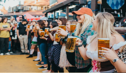 A group of people holding large beers, smiling and celebrating at an outdoor event. Festive atmosphere with colorful decorations.