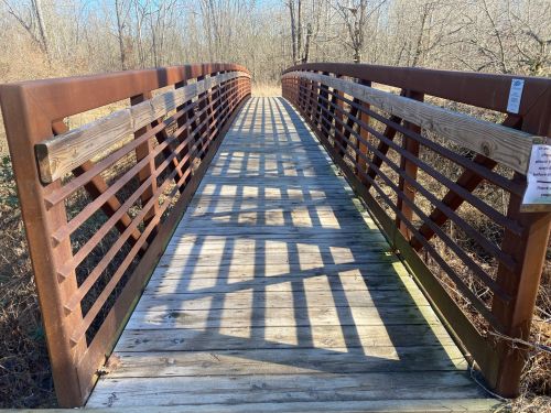 A wooden bridge with metal railings spans a path through a wooded area, casting shadows on the ground.