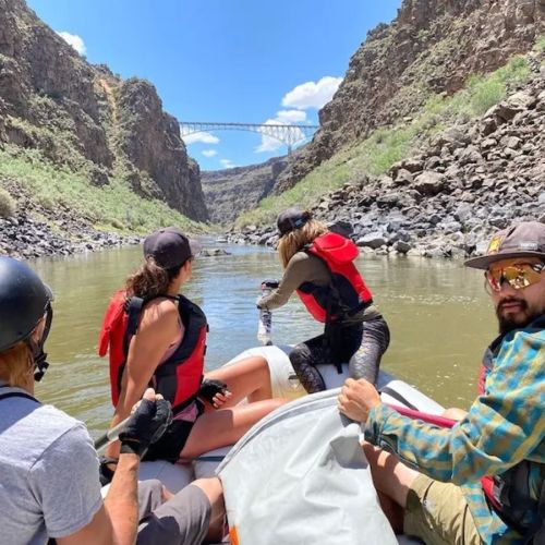 A group of people in a raft navigating a river surrounded by rocky cliffs and a bridge in the background under a blue sky.