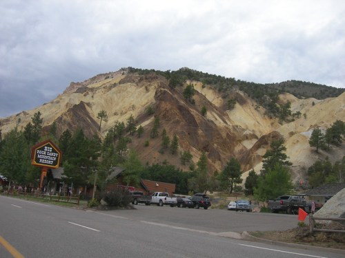 A scenic view of colorful rock formations and pine trees near a roadside attraction. Cloudy sky above.
