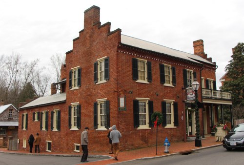 Historic brick building with black shutters, surrounded by trees, and people walking nearby on a cloudy day.