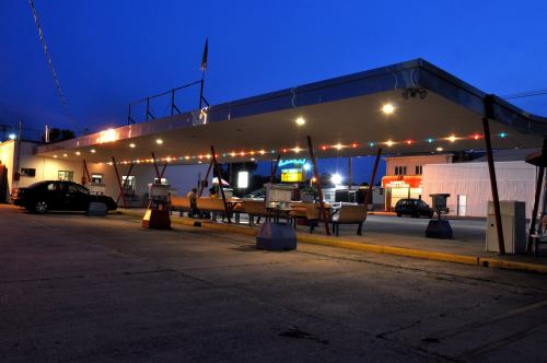 A retro diner with outdoor seating, illuminated by colorful lights, at dusk. Cars are parked nearby.