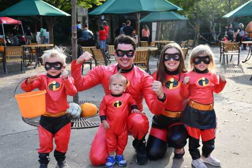 A family dressed as superheroes poses together, flexing their muscles in matching red costumes with black masks.