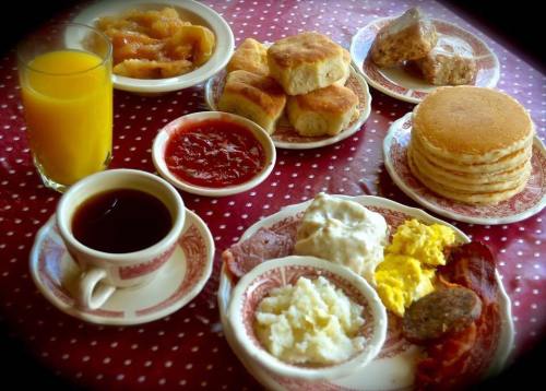 A breakfast spread featuring pancakes, biscuits, eggs, bacon, sausage, grits, fruit preserves, and orange juice.