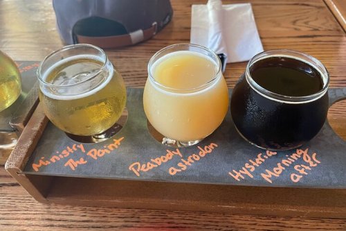 A wooden flight tray with three glasses of beer: a light beer, a hazy yellow beer, and a dark stout.