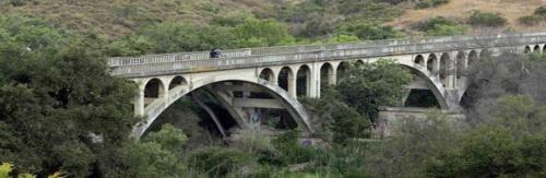 A concrete arch bridge spans a lush green landscape, surrounded by trees and hills.